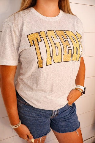 A woman wearing a light heather gray t-shirt with 'TIGERS' printed across the chest in a gold glitter-textured font, paired with denim shorts.