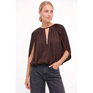 A woman wearing a dark brown draped blouse with a keyhole cutout and wide cap sleeves, paired with dark gray jeans, against a plain white studio background.