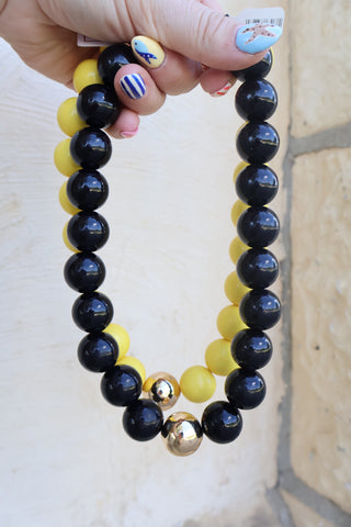 A hand holds a beaded necklace featuring glossy black and matte yellow round beads, with two small metallic gold accent beads at the bottom, set against a light beige stone wall background.