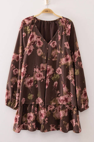A long-sleeve brown blouse with a pink and green floral print, featuring a split V-neckline and a ruffled hem, hanging on a wooden hanger against a plain light gray wall.