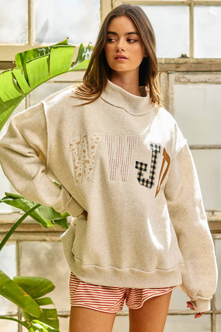 Woman wearing a beige sweatshirt with text and graphics, standing indoors with plants in the background