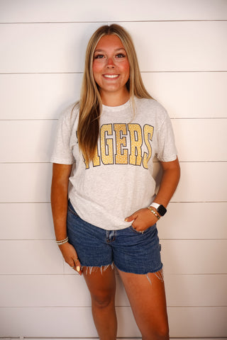 A smiling young woman stands against a white shiplap wall wearing a light heather gray graphic t-shirt with 'TIGERS' printed in gold across the chest, paired with denim cutoff shorts.