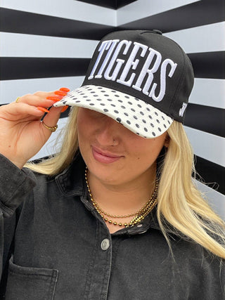 Person wearing a black cap with 'TIGERS' text and a polka dot visor against a striped background