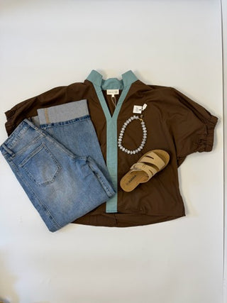 A styled flat-lay of a women's brown short-sleeve top with a light blue collar and front placket, paired with folded blue jeans, a pearl necklace, and tan slide sandals on a white background.