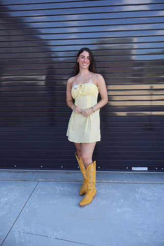 A woman stands in front of a dark gray metal roll-up door wearing a pale yellow strapless mini dress and tall mustard yellow cowboy boots.