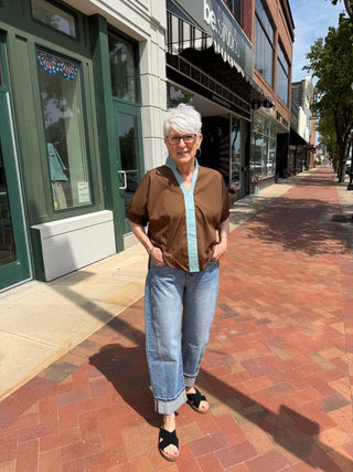 An older woman with short white hair stands on a brick sidewalk in front of storefronts, wearing a brown short-sleeve button-up shirt with a light blue striped collar and placket, light blue jeans, and black slide sandals.