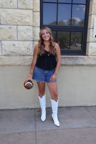 A young woman stands outdoors in front of a stone building wall and window, wearing a black sleeveless top, denim shorts, white cowboy boots, and holding a football.