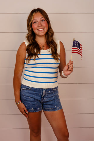 Woman in a striped tank top and denim shorts holding an American flag against a wooden background