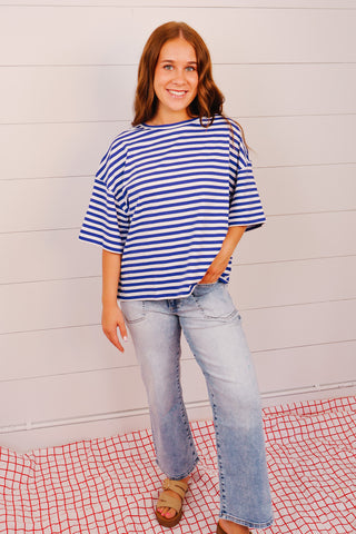 Woman wearing a blue and white striped shirt and jeans standing against a light wooden panel background.