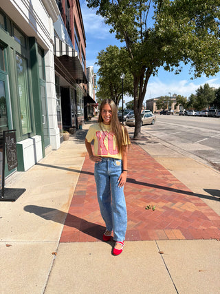 A young woman stands on a city sidewalk wearing a beige graphic t-shirt with pink and yellow lettering, light blue wide-leg jeans, and red sneakers. The background shows a street with parked cars, trees, and storefronts under a blue sky.