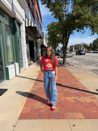 A young woman stands on a brick sidewalk in front of storefronts and trees, wearing a red graphic T-shirt, light blue wide-leg jeans, and red sneakers.