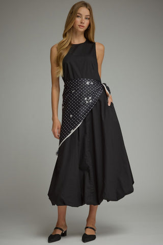 A woman modeling a sleeveless black midi dress with a wide, draped waist panel covered in white polka dots and silver floral appliqués, paired with black pointed flats against a neutral gray studio background.