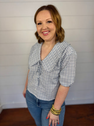 Woman wearing a blue checkered blouse and jeans against a white wall.