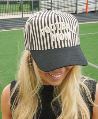 A woman on a sports field wearing a black and white vertically striped baseball cap with 'FOOTBALL MOM' embroidered in white on the front.