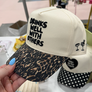 A hand holds a beige baseball cap with a leopard-print brim and black embroidered text reading 'DRINKS WELL WITH OTHERS.' The cap also has small black cocktail graphics on the side.