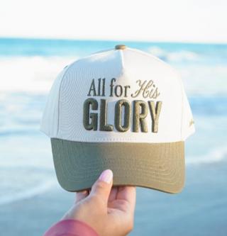 A hand holds a white and olive green trucker hat with the text 'All for His GLORY' embroidered on the front, set against a blurred beach background.