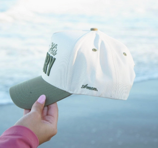 A hand holds a white and sage green baseball cap with 'Hershey's Pasty' embroidered on the front, set against a blurred beach background.