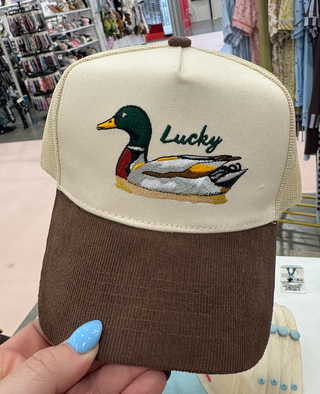 A close-up of a trucker-style cap being held by a hand, featuring a cream front panel with an embroidered mallard duck and the word 'Lucky,' and a brown corduroy brim. The mesh back is tan.