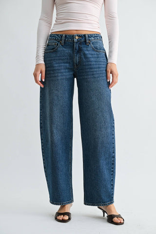 A model wearing a pair of high-waisted, wide-leg blue denim jeans, paired with a light pink long-sleeve crop top and dark brown heeled sandals, photographed against a plain light gray studio background.
