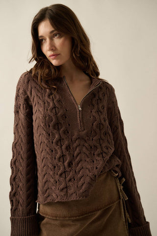 A woman wearing a brown cable-knit half-zip sweater with a ribbed collar, cuffs, and hem, paired with a brown skirt, set against a plain beige studio background.