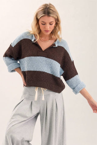 A woman wearing a chunky knit polo-style sweater with horizontal brown and light blue stripes, paired with light gray drawstring pants, against a plain light background.