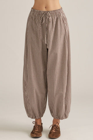 A person wearing loose, high-waisted checkered pants with a drawstring waist and elastic cuffs, paired with brown lace-up shoes, against a plain light beige studio background.