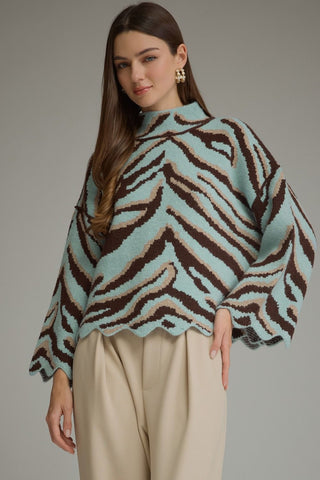 A woman modeling a fuzzy knit mock-neck poncho sweater with a brown, beige, and mint green zebra-like pattern, paired with beige trousers, against a solid gray studio background.